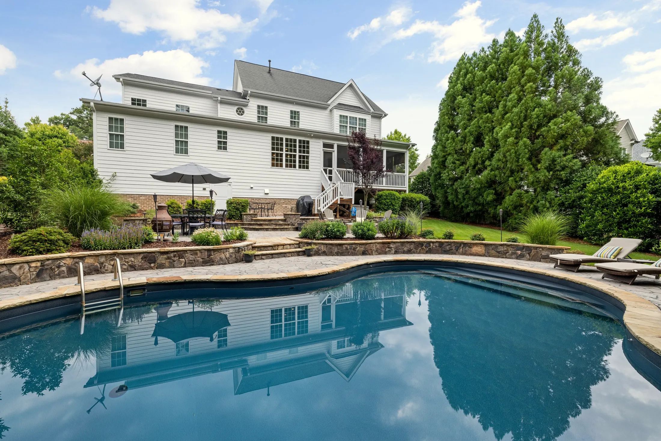 Backyard swimming pool with stone coping, with the back of the white house and trees beyond