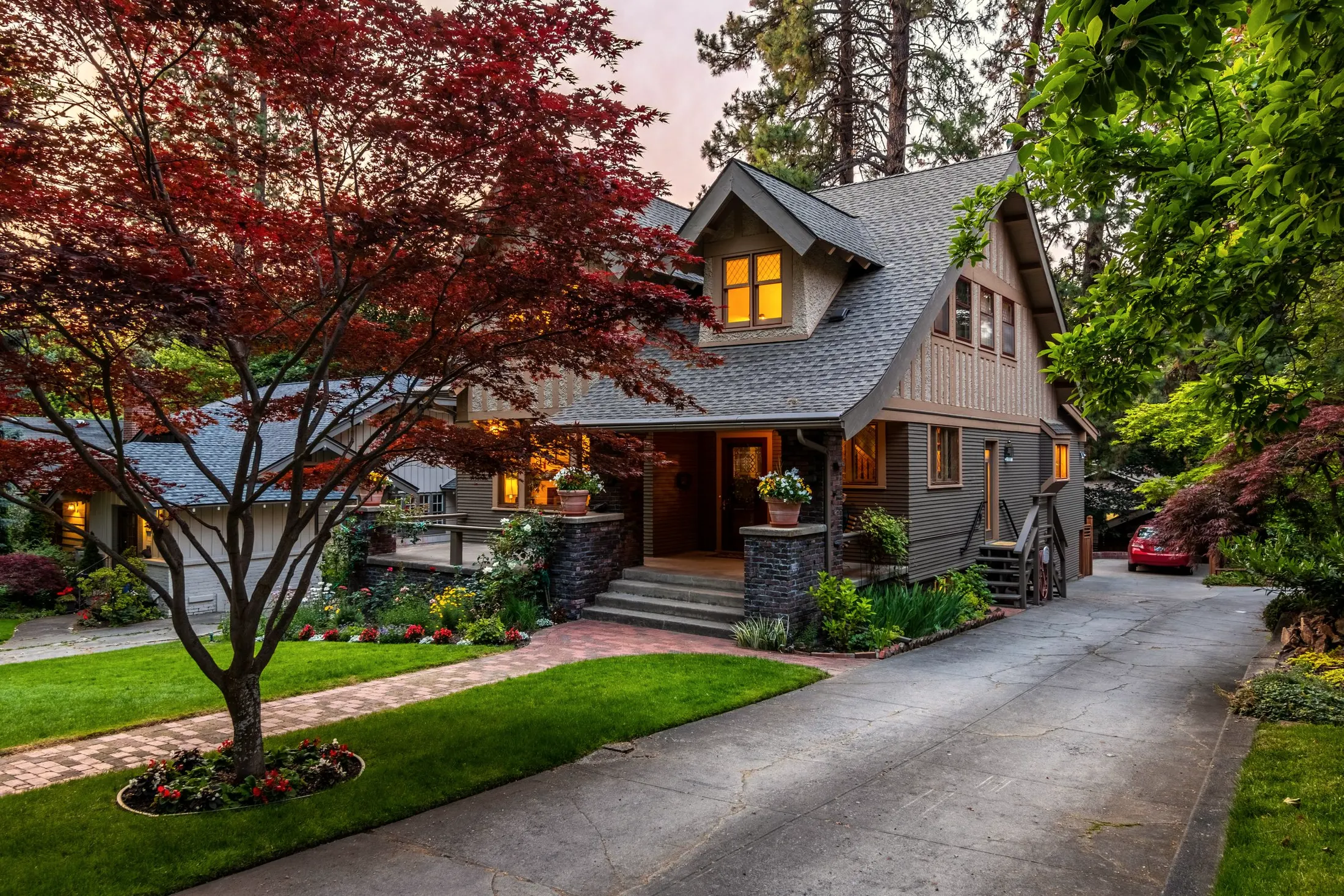 A craftsman house in the evening, its lit windows glowing above a lawn, with a paved drive curving past a red maple