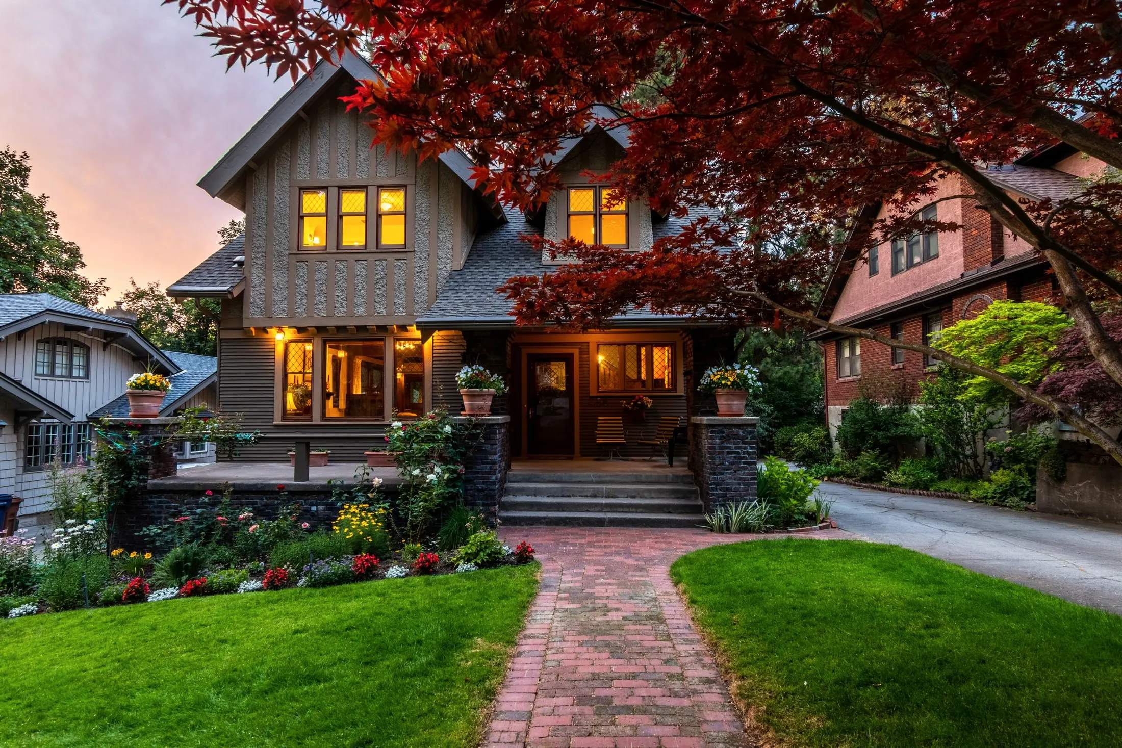 A craftsman-style house at dusk with warm lit windows, a brick path and a red maple on the lawn