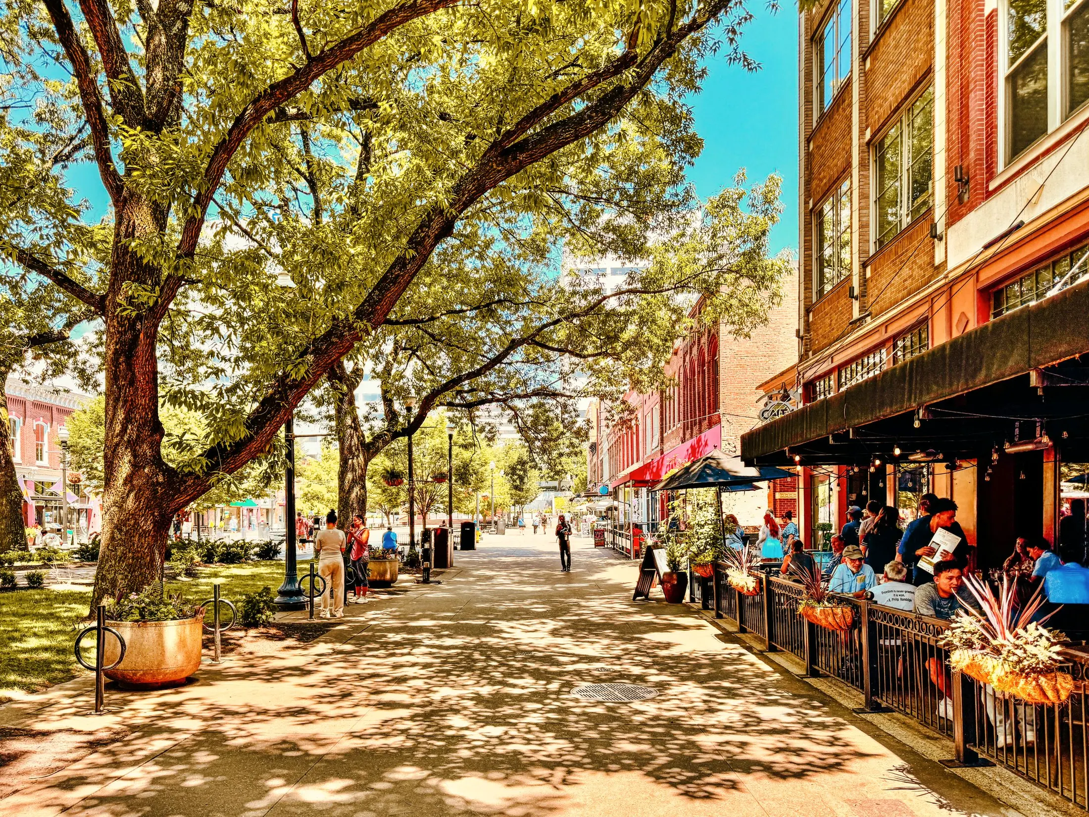A tree-shaded street with restaurant patios and people walking on the sidewalk
