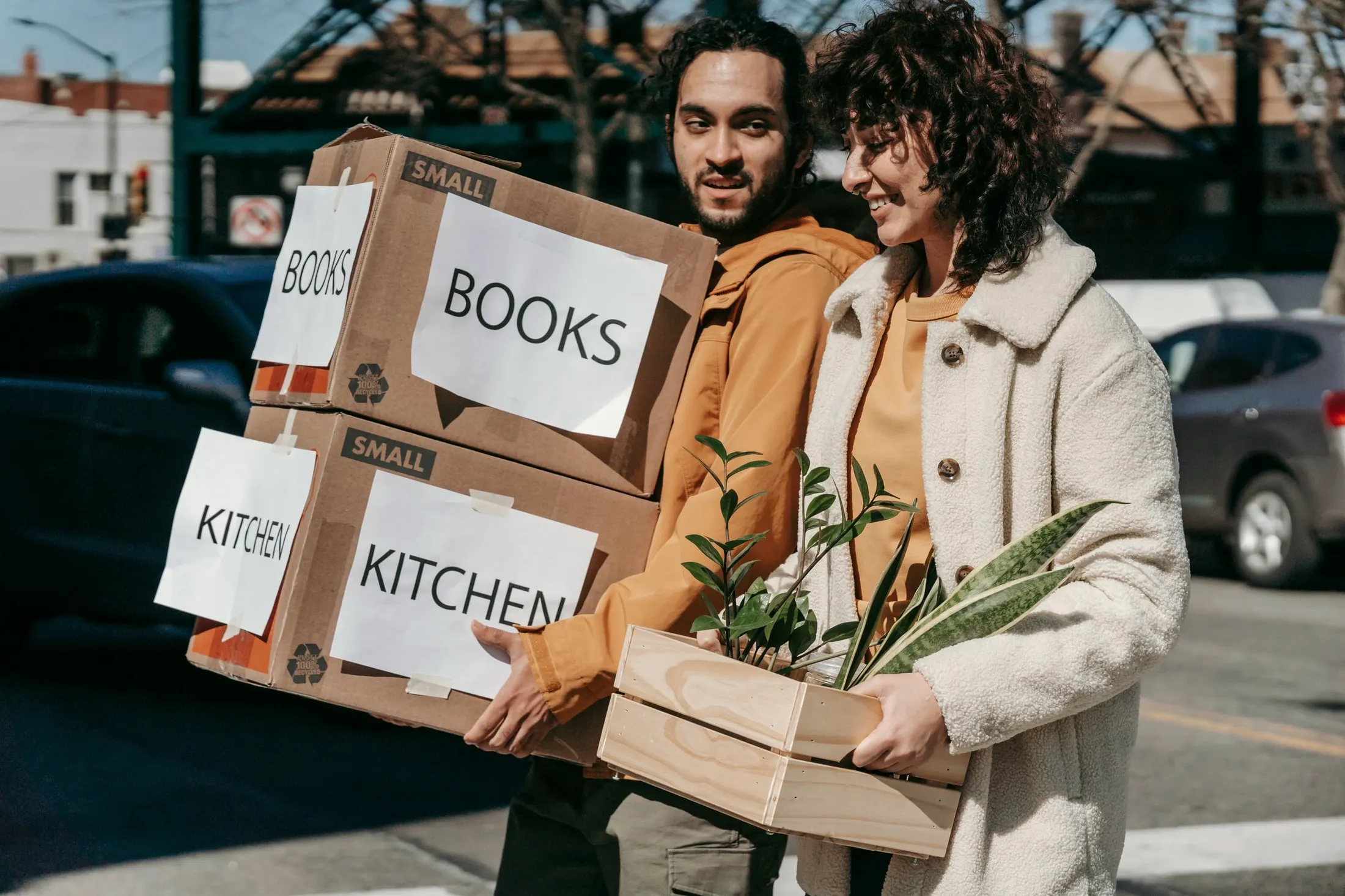 A couple carrying moving boxes labeled books and kitchen, with a plant, along a city sidewalk