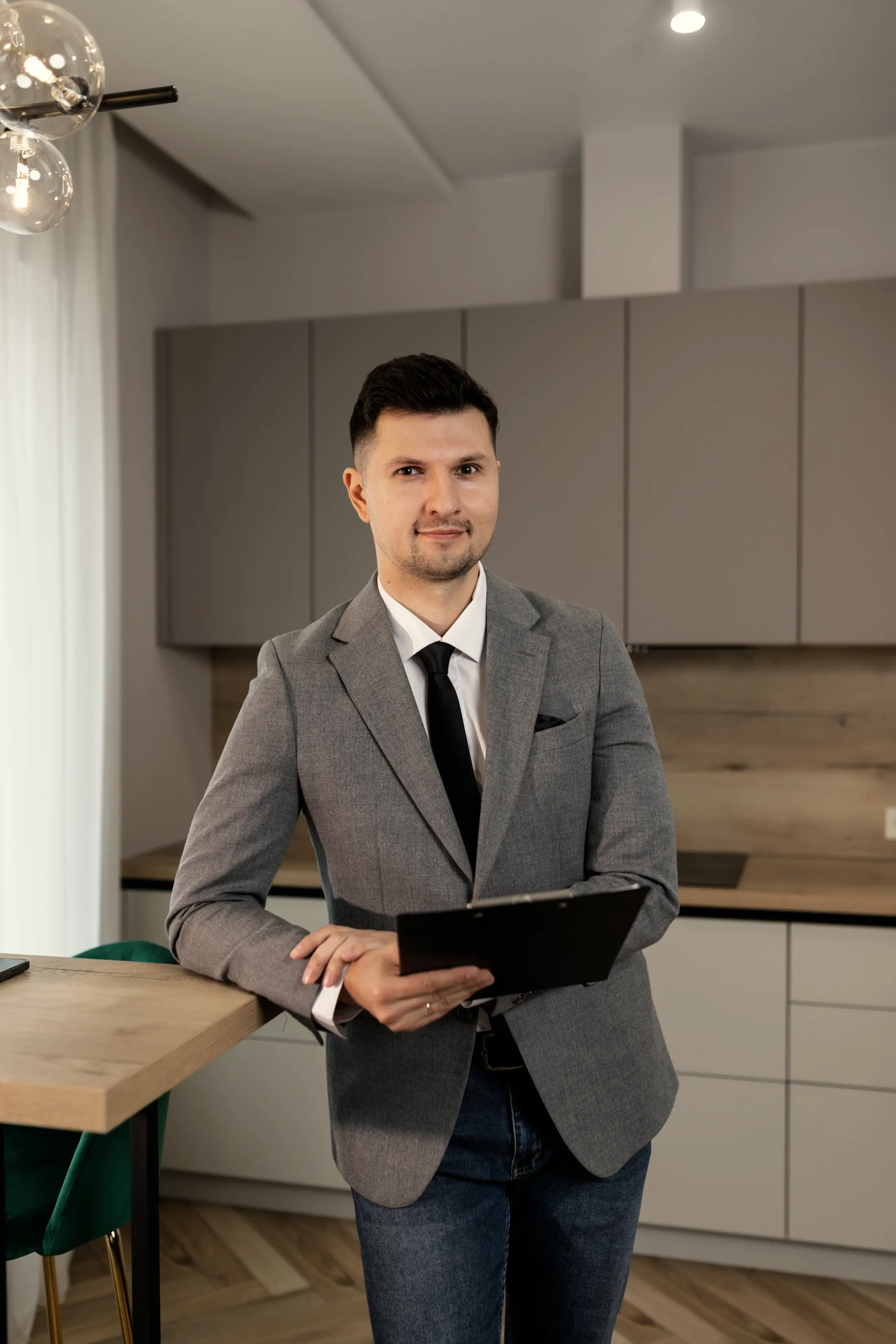 Portrait of a man in a gray suit jacket holding a folder, standing in a bright kitchen
