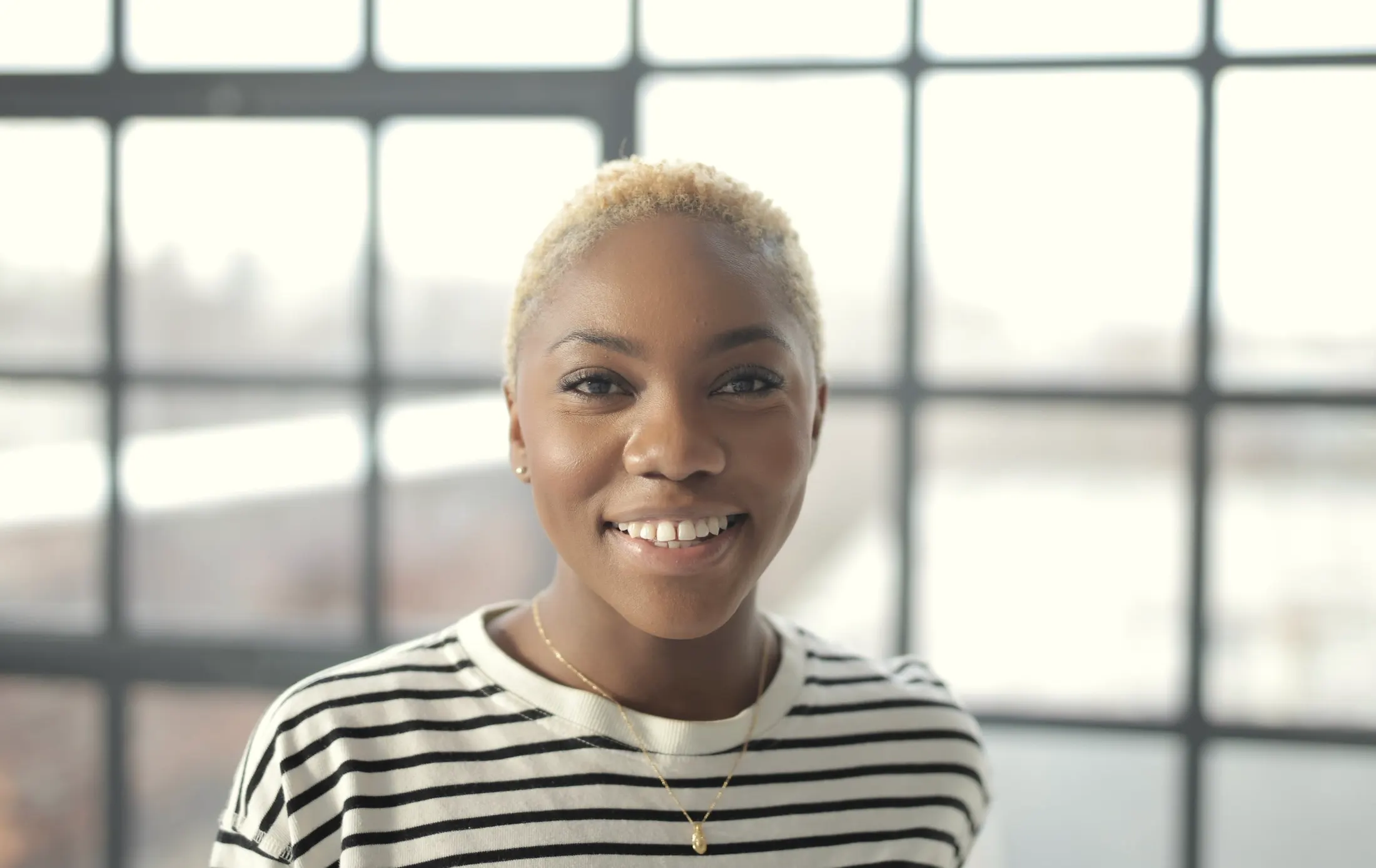 Portrait of a smiling woman with short blond hair in a striped shirt, in front of tall windows