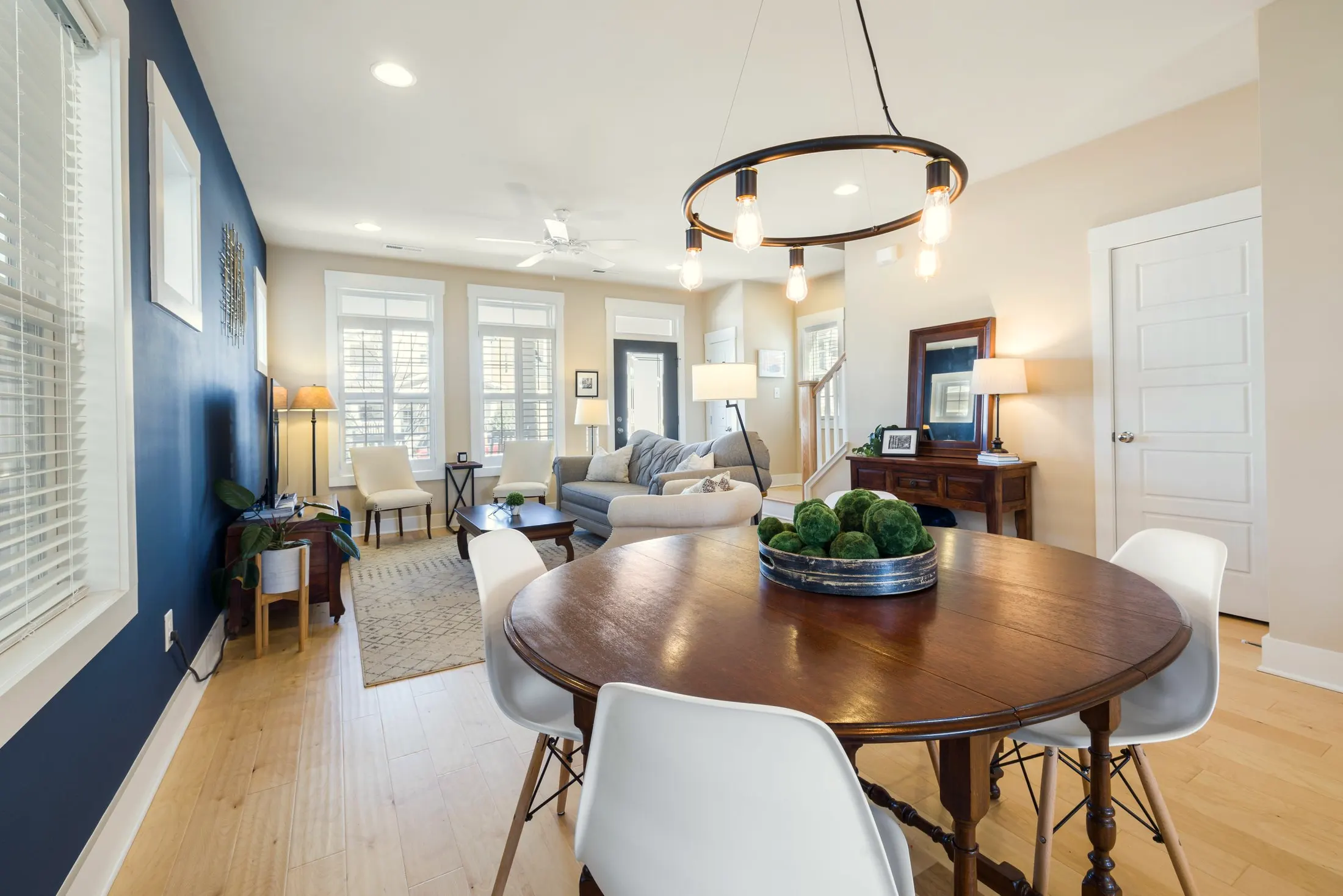Round dining table with white chairs beside a navy wall, with a sitting area and a ring chandelier beyond